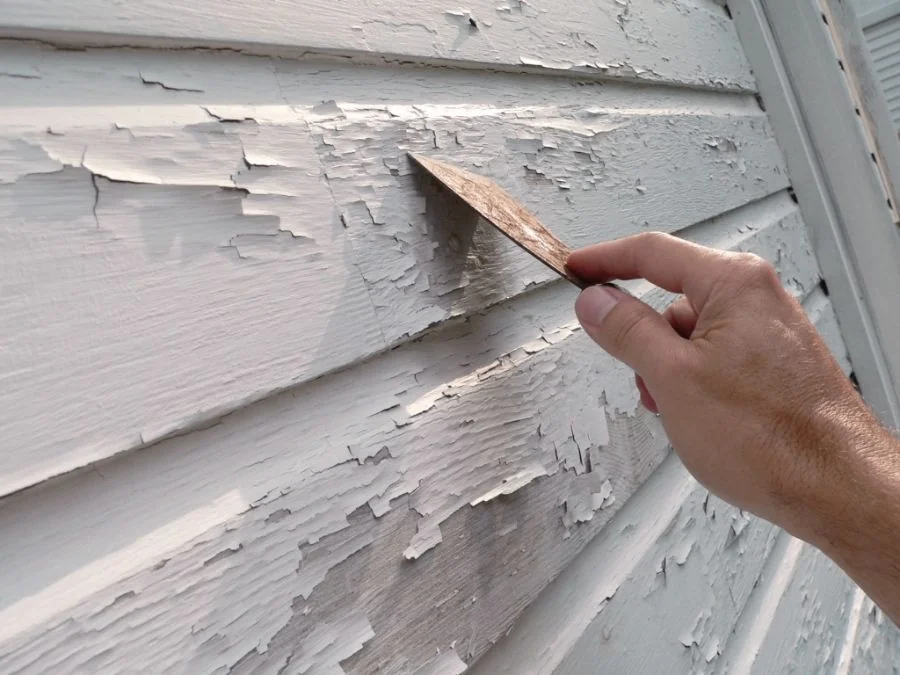 A hand scraping peeling paint off exterior wood siding with a putty knife.